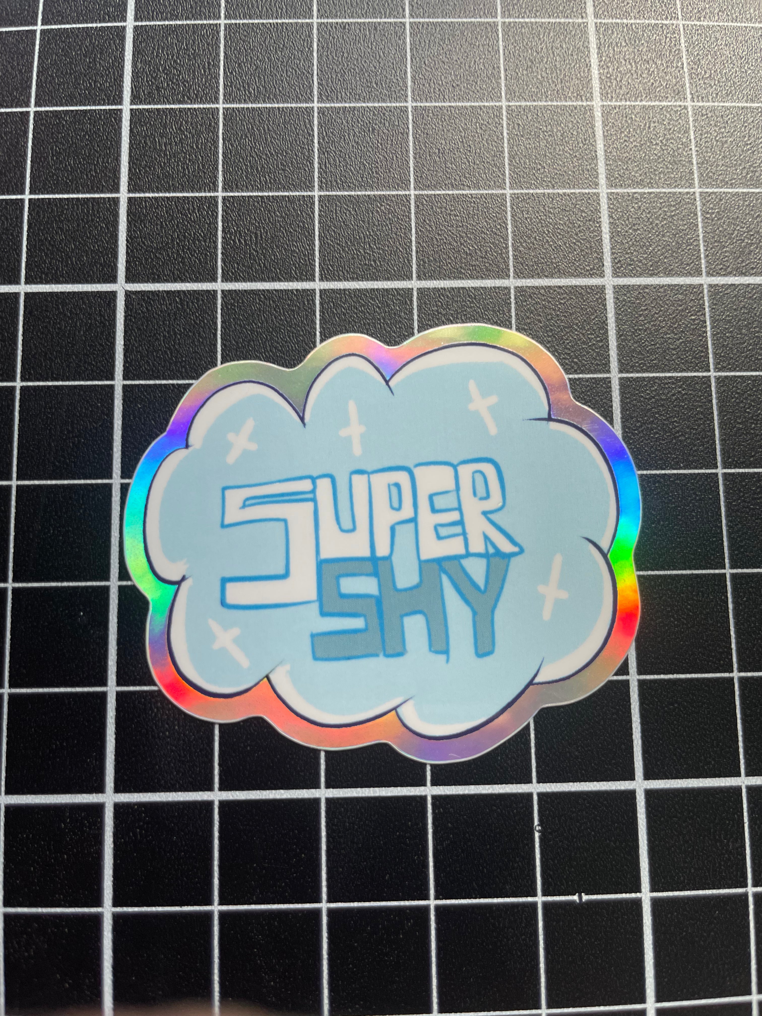 Holographic 'Super Shy' sticker on a grid background