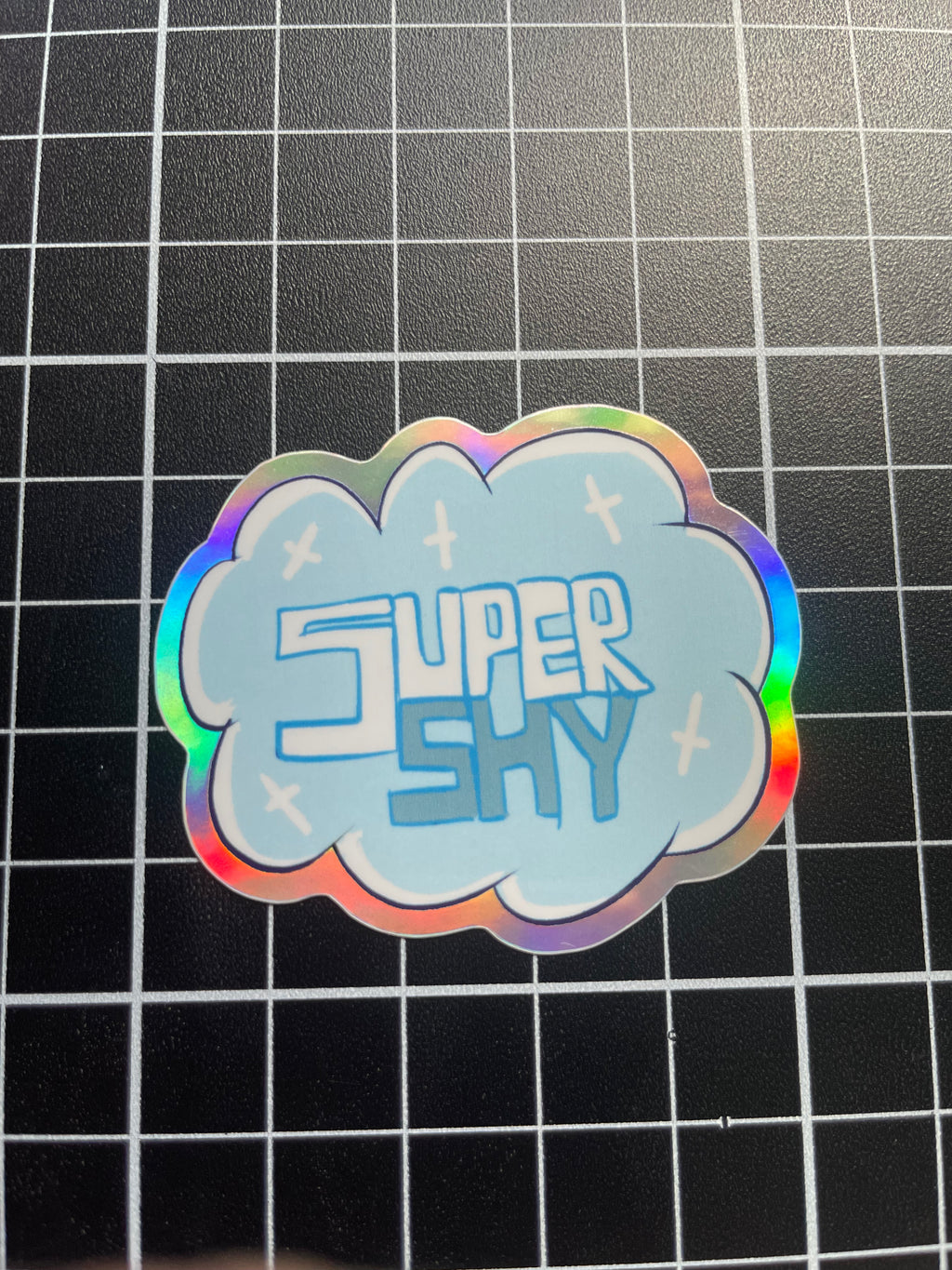 Holographic 'Super Shy' sticker on a grid background